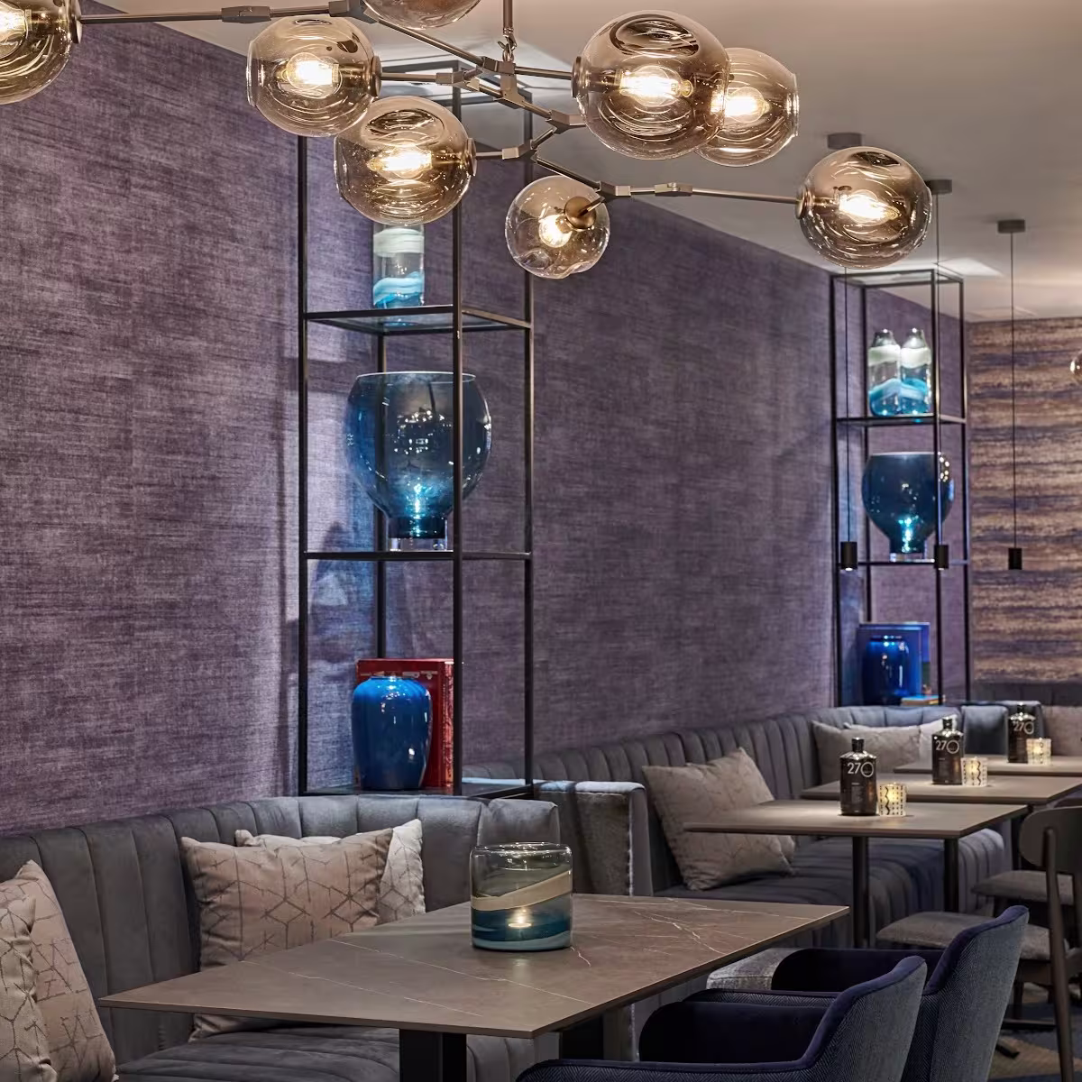 Espacio elegante con sofás grises, sillas tapizadas y una lámpara moderna de esferas. Decoración en tonos azules con jarrones y velas sobre las mesas.