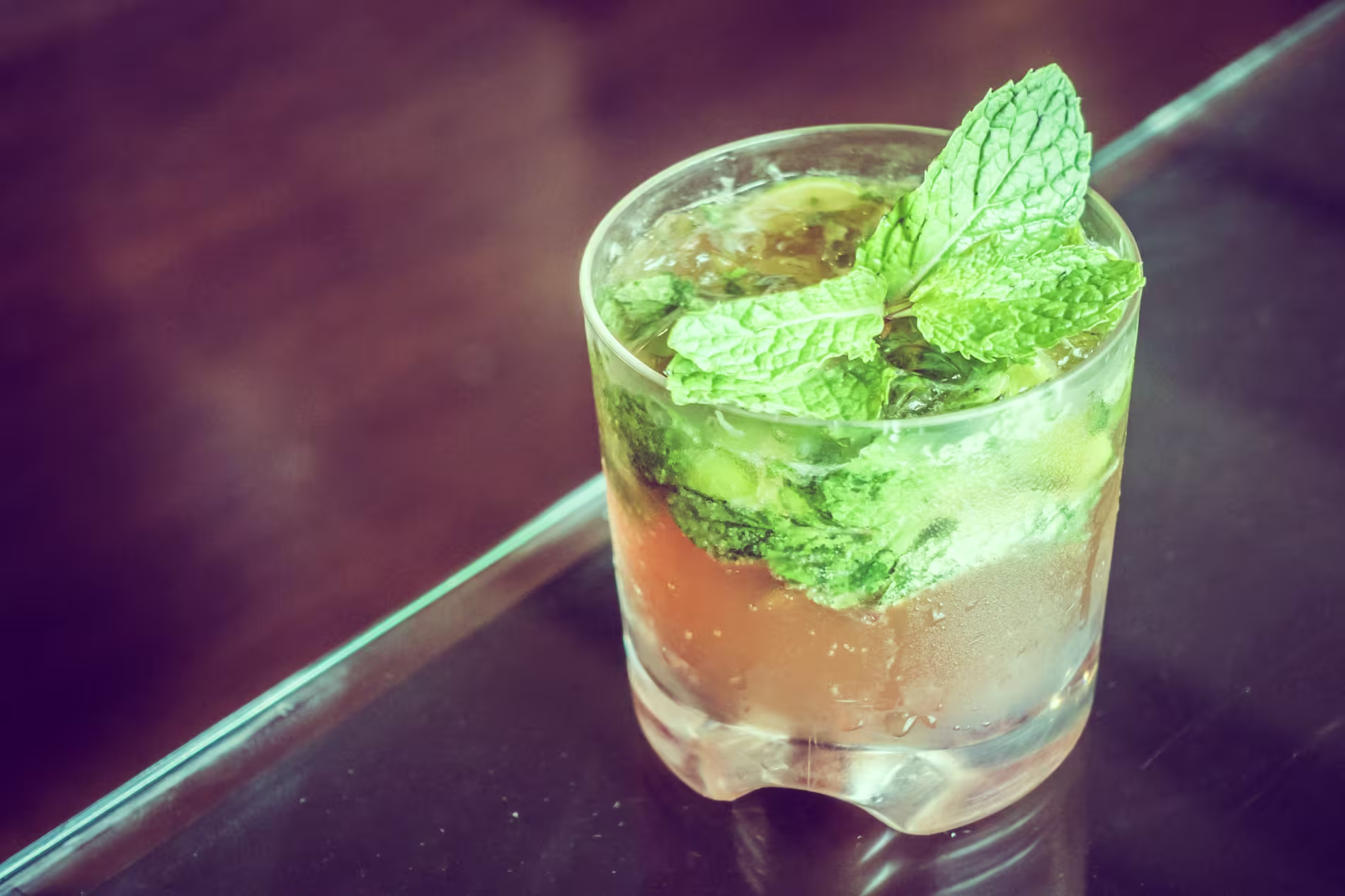 A glass of mojito cocktail with ice, mint leaves, and lime, placed on a reflective surface.