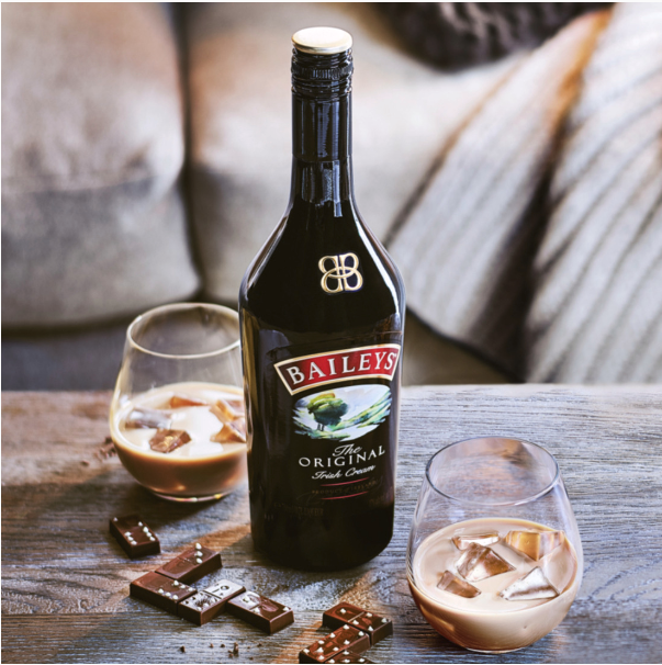 Botella de Baileys con dos vasos , hielo y la bebida en cada uno. Chocolate en la mesa y sillon detrás.