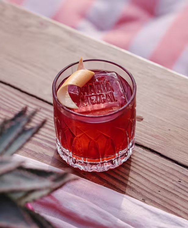 Trago en mesa de madera. El trago es color rojo, con un gran hielo grabado maria mezcal y una rodaja de limón como decoración