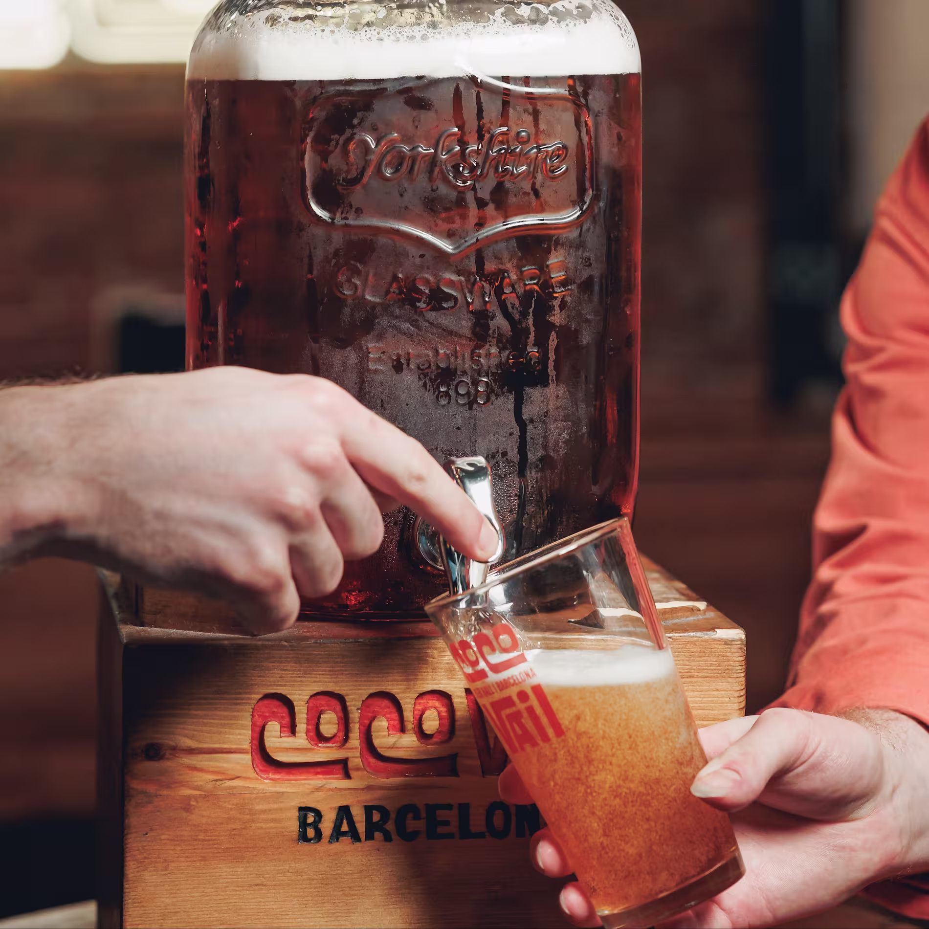 La imagen muestra las manos de una persona sirviendo una bebida de un gran tarro de vidrio con un grifo en un vaso más pequeño. El tarro está etiquetado con la palabra “Cocktails” y tiene “Barcelona” escrito debajo. 