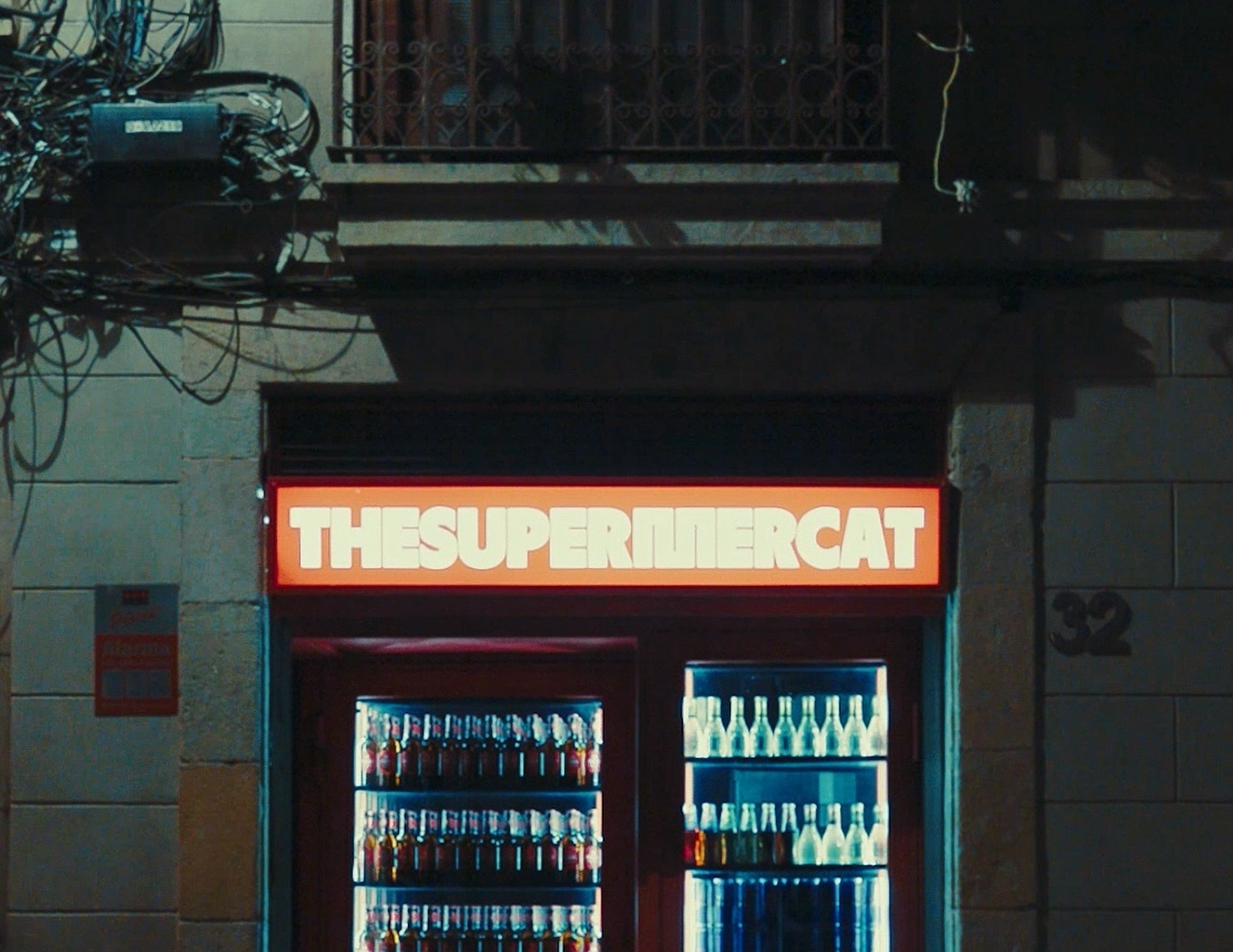 Fachada de The Supermercat iluminada de noche, con neveras repletas de bebidas destacando detrás de un cartel vibrante y moderno.