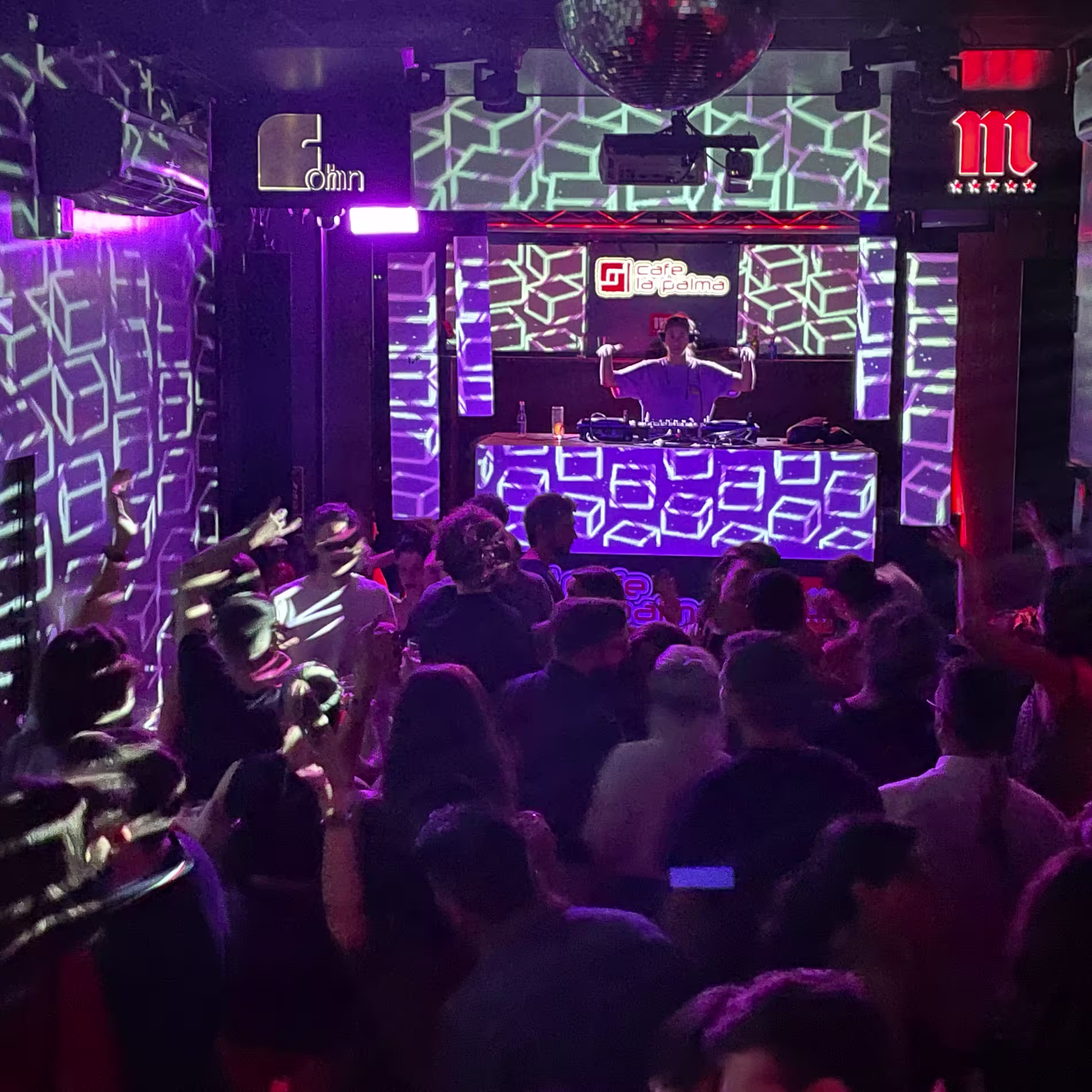 Pista llena y luces geométricas en el corazón de Café La Palma, con el DJ animando a la multitud.