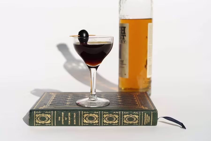 A cocktail garnished with a cherry sits on a closed book, with a partially visible bottle of whiskey in the background.