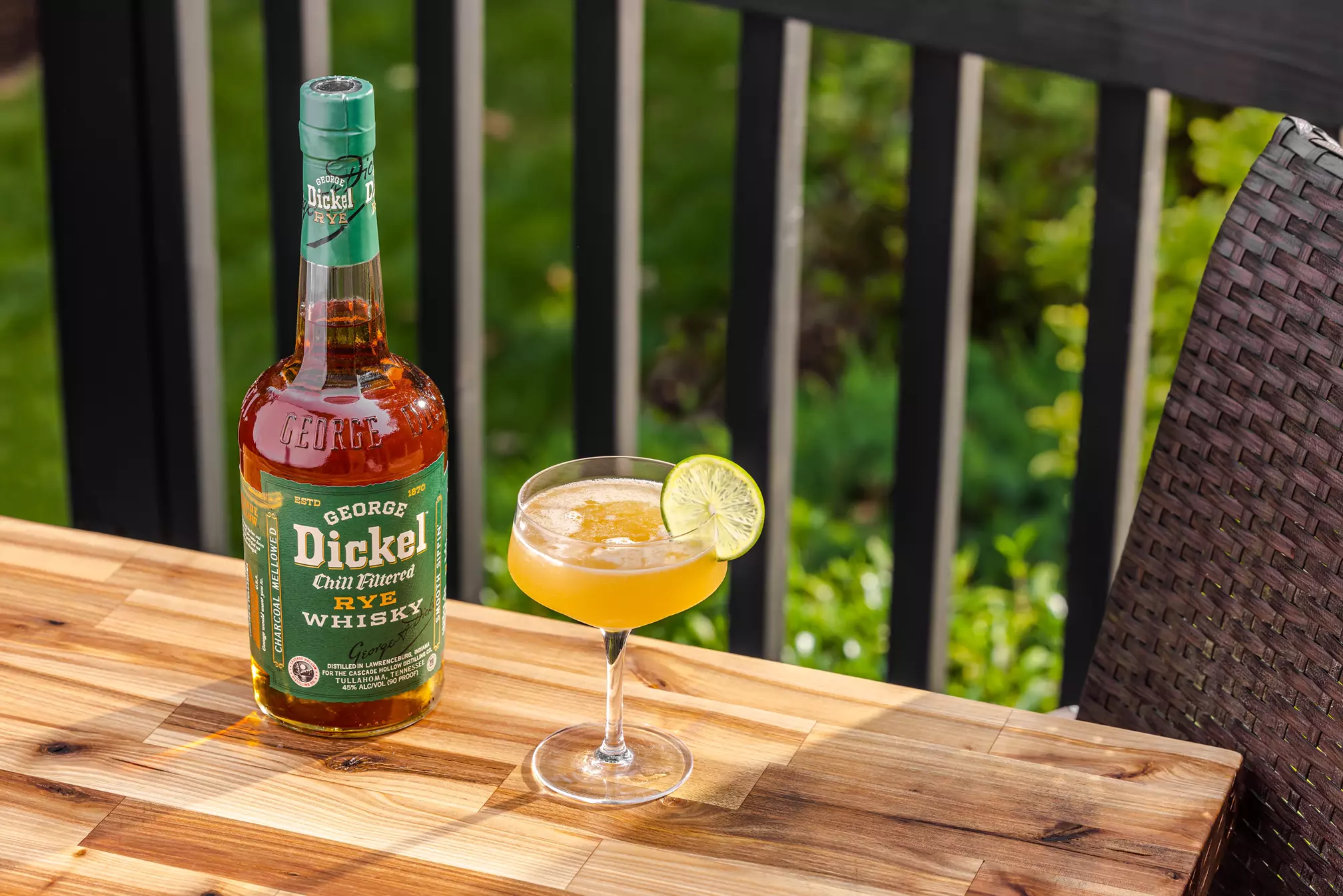 A bottle of George Dickel Rye Whisky sits next to a cocktail garnished with a lime wheel on a wooden table outdoors.