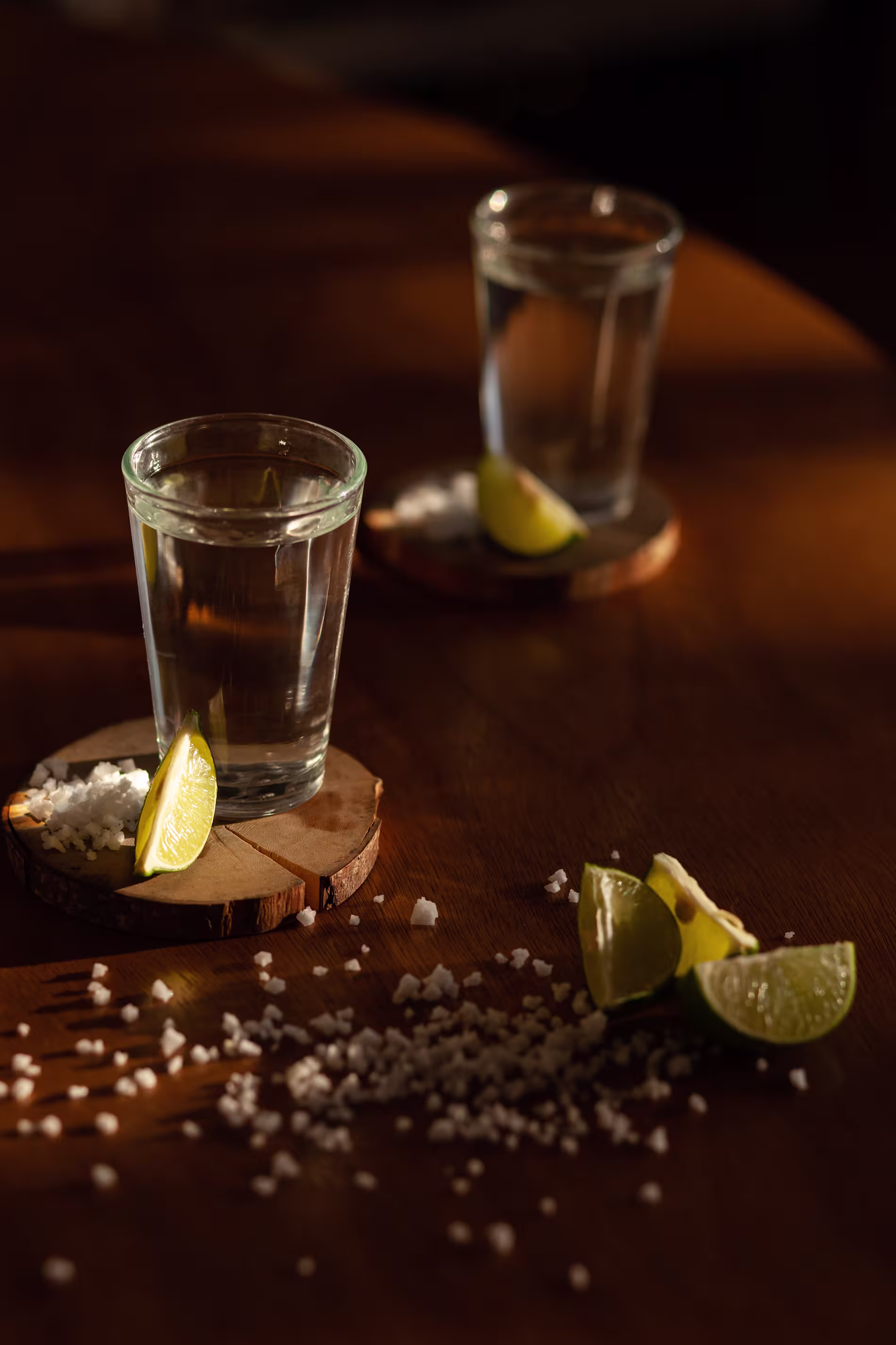 Two glasses of clear liquid on coasters, each with a lime wedge. Coarse salt and additional lime wedges are scattered on the wooden surface.
