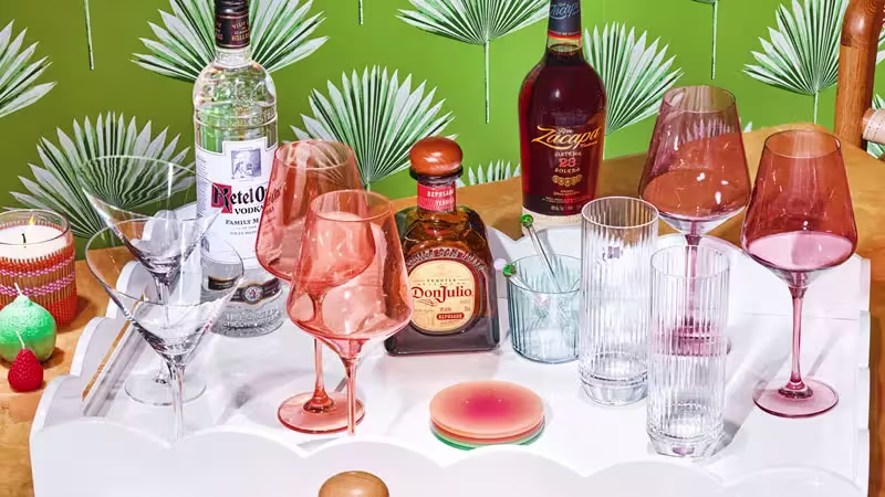 A white tray holding assorted liquor bottles, drinking glasses, and cocktail glasses sits on a table with a green leafy-patterned background.