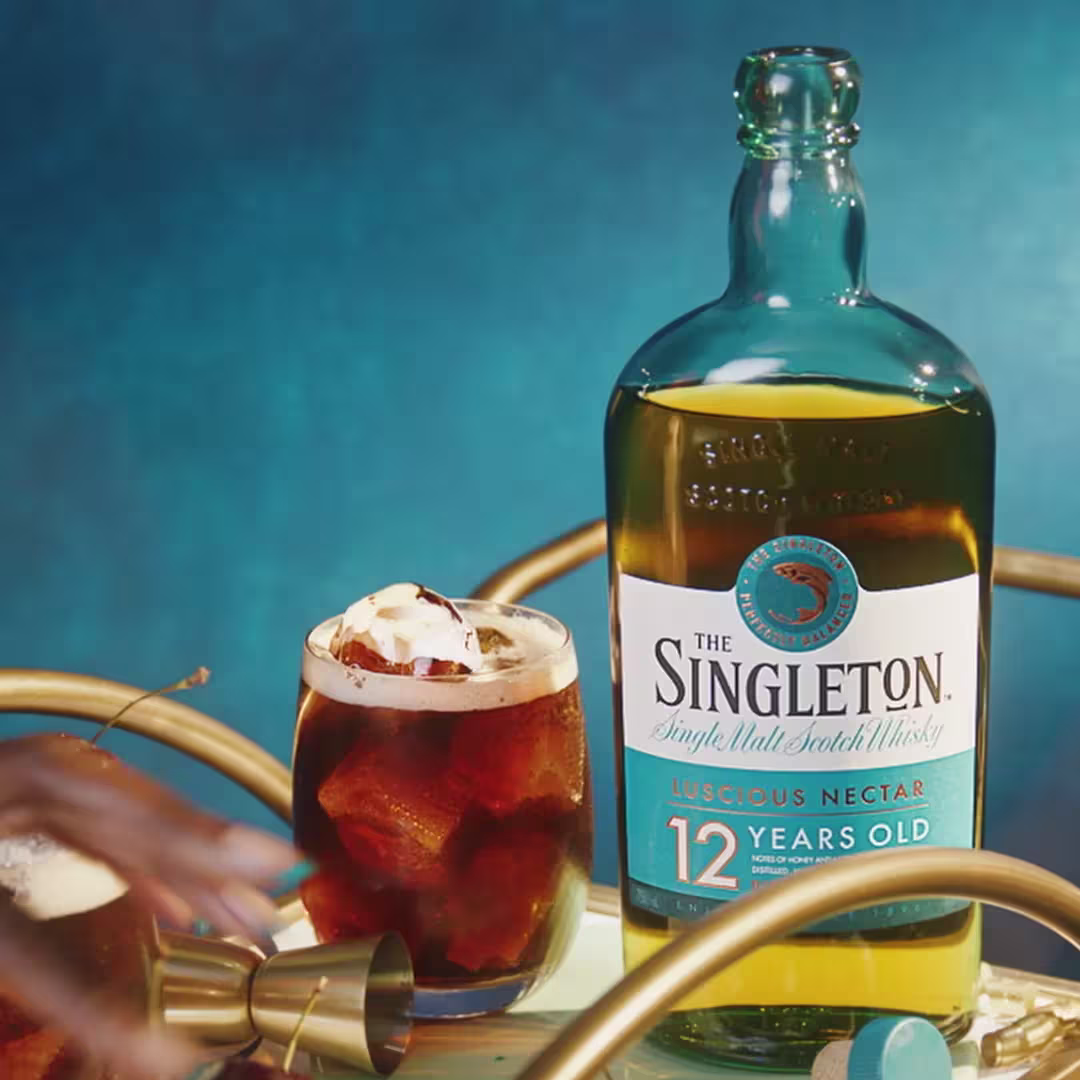 A bottle of The Singleton 12-year-old single malt Scotch whisky next to a glass of iced cocktail on a gold bar cart against a blue background.