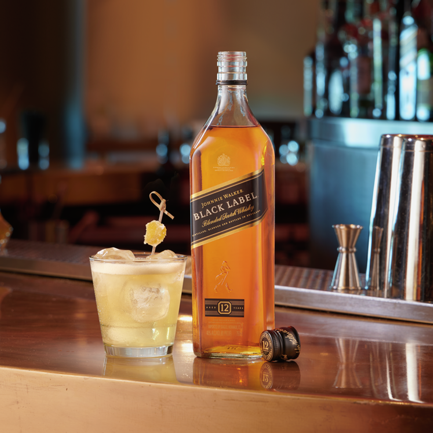 En la imagen se puede observar un vaso con el cóctel Penicillin y, al lado, una botella de Whisky Johnnie Walker Black Label, apoyados sobre una barra de bar.