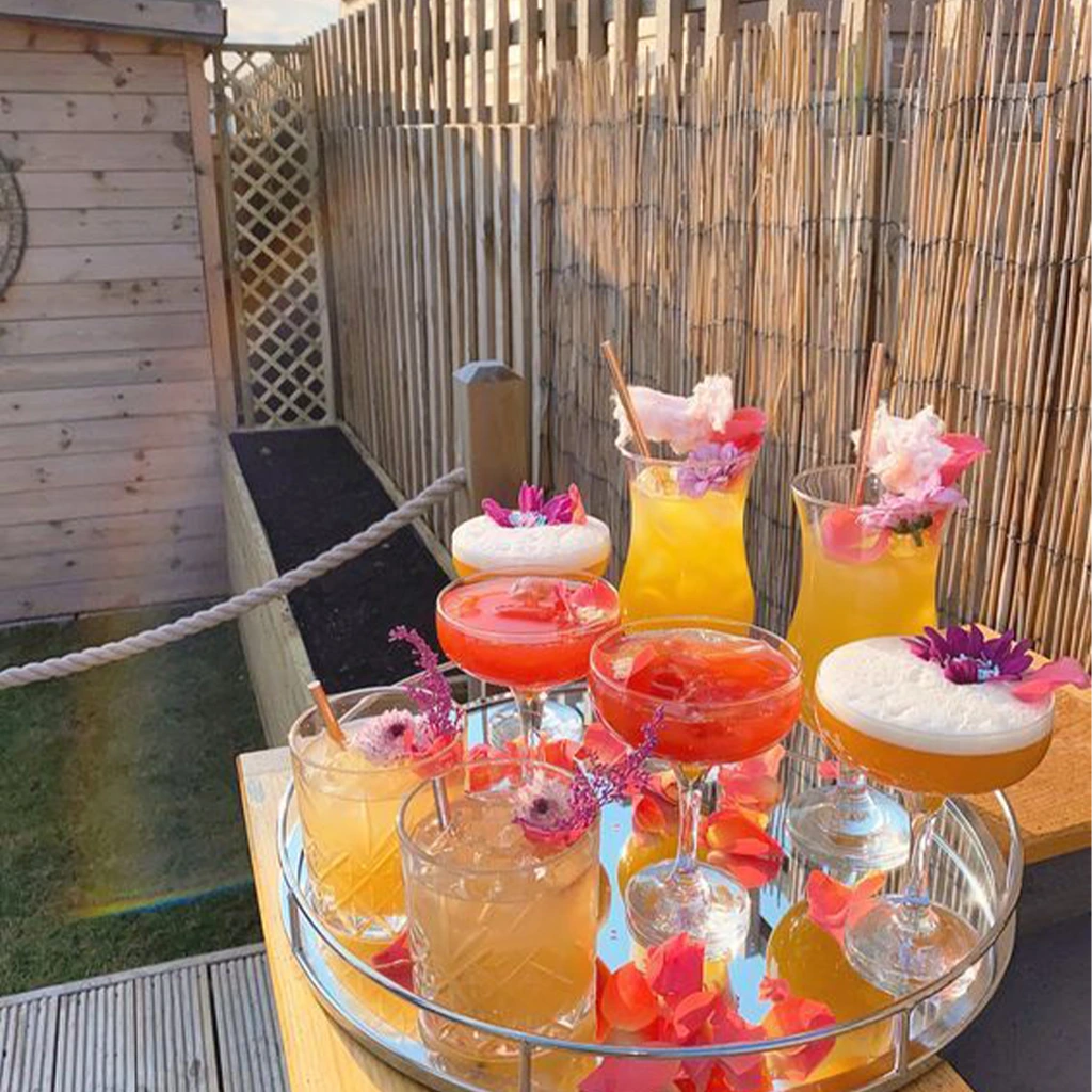 A tray with eight colorful cocktails garnished with flowers and fruit sits on an outdoor table near a wooden fence and shed.