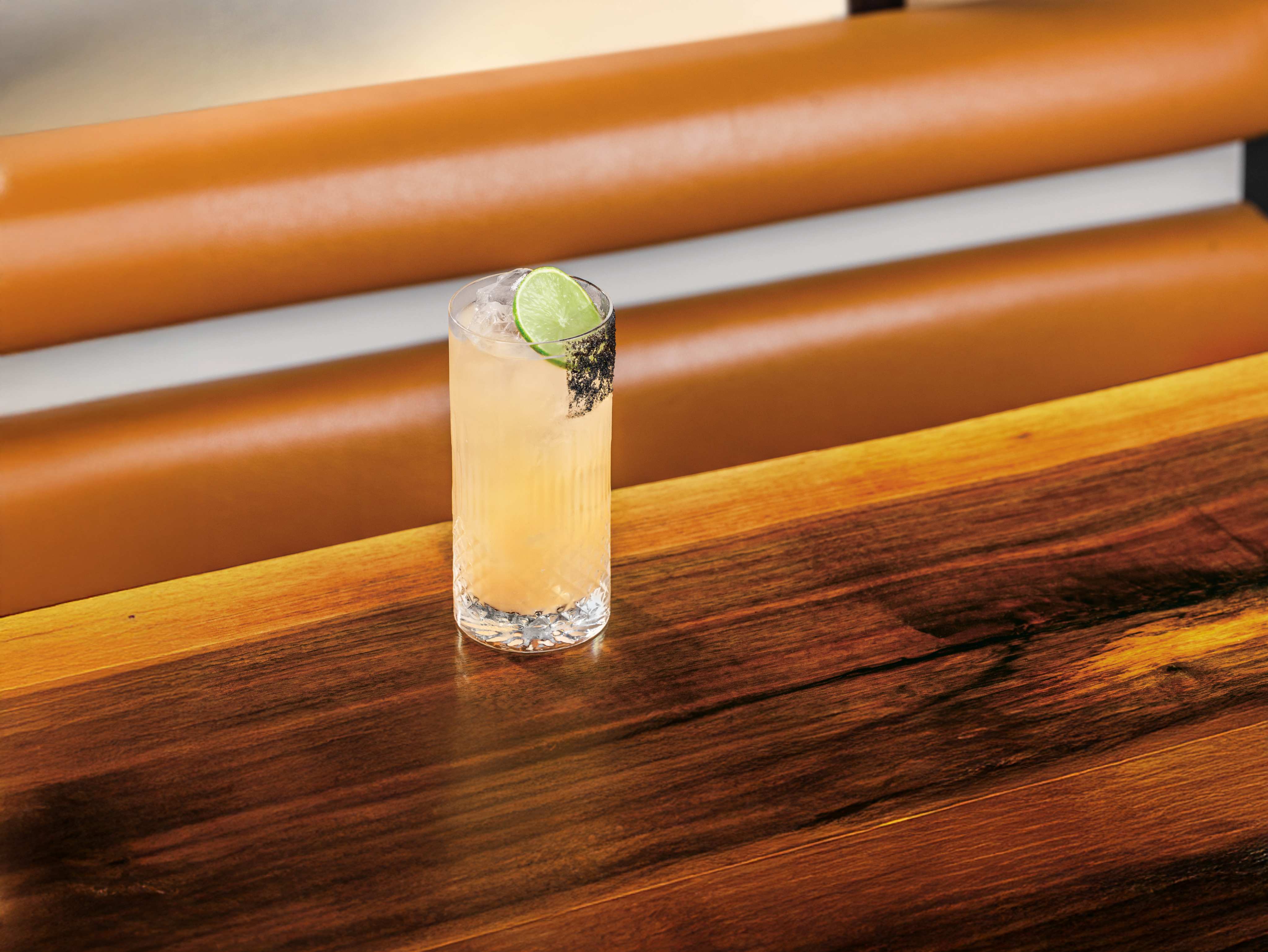 A tall glass of light-colored cocktail with ice and a lime wedge sits on a polished wooden table in front of a tan cushioned bench.