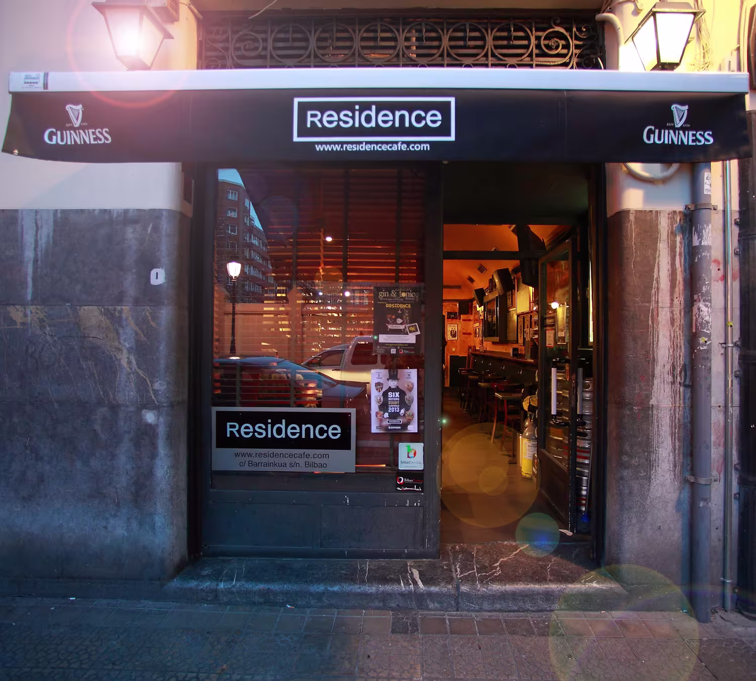 Fachada de Pub Residence en Bilbao, con toldo negro de Guinness, interior cálido con barra de madera y carteles promocionales en la entrada.