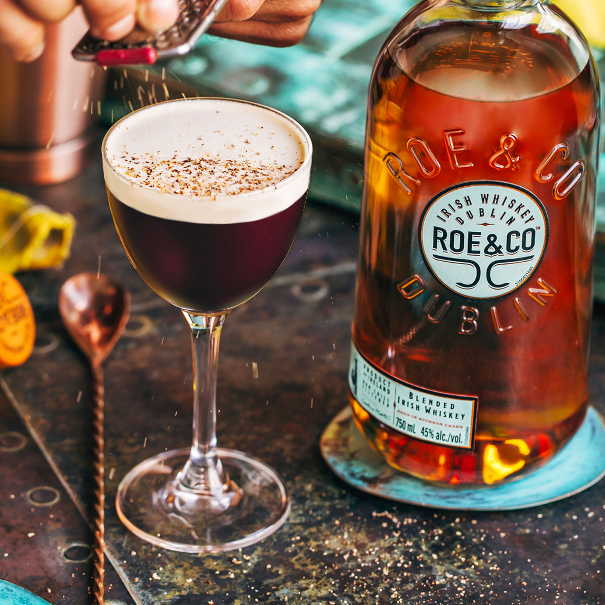 A glass of Irish coffee with foam is sprinkled with grated chocolate next to a bottle of Roe & Co Irish whiskey on a rustic table.