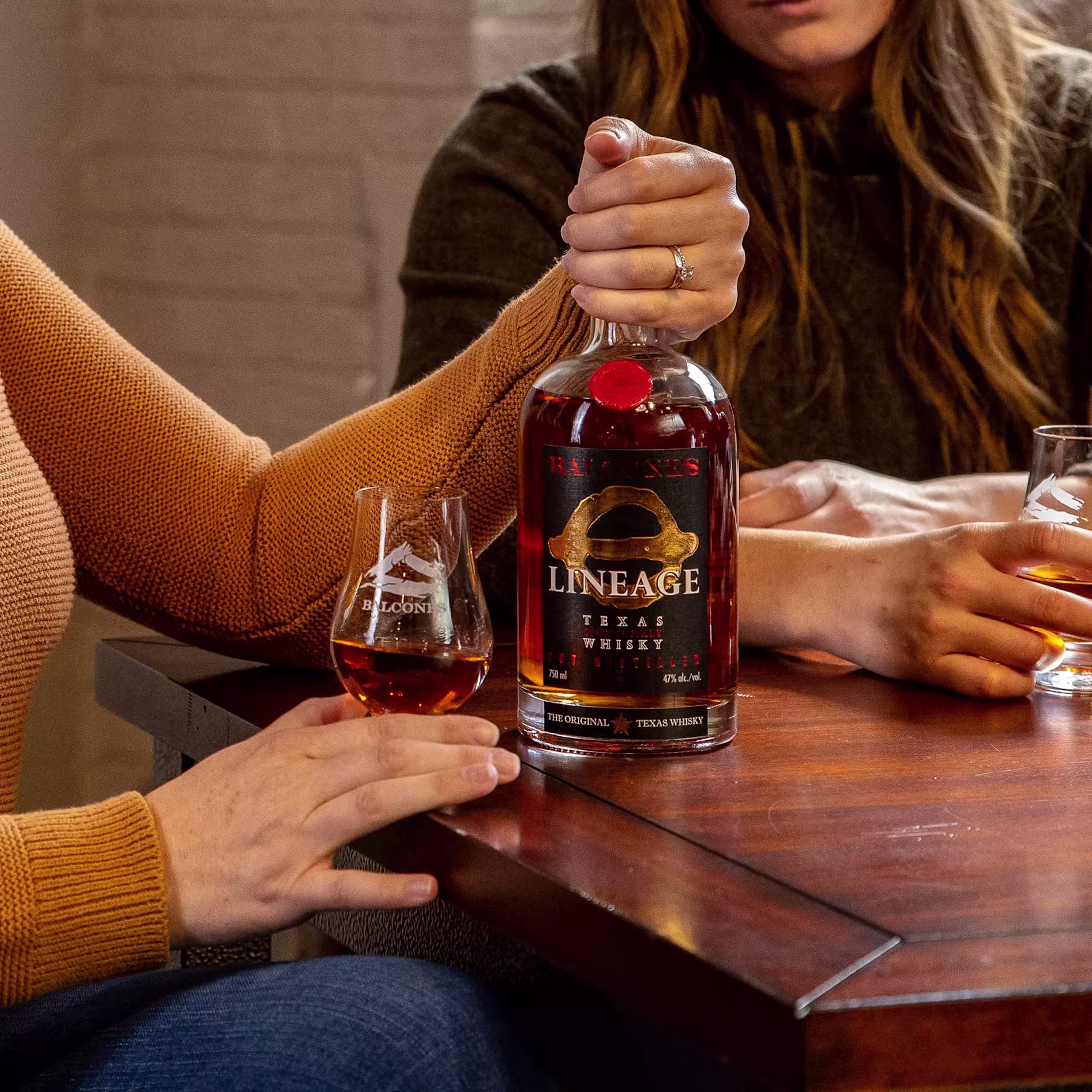 Two people sit at a wooden table with a bottle of Balcones Lineage Texas Single Malt Whisky and two glasses of whisky.