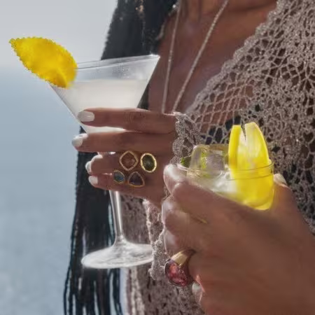 Dos manos sostienen cócteles; un vaso tiene una guarnición de limón, el otro un cóctel amarillo con hielo. La persona lleva anillos y un top de ganchillo, con el océano visible al fondo.
