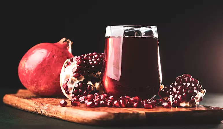 A glass of pomegranate juice sits on a wooden board surrounded by a whole pomegranate and scattered pomegranate seeds against a dark background.