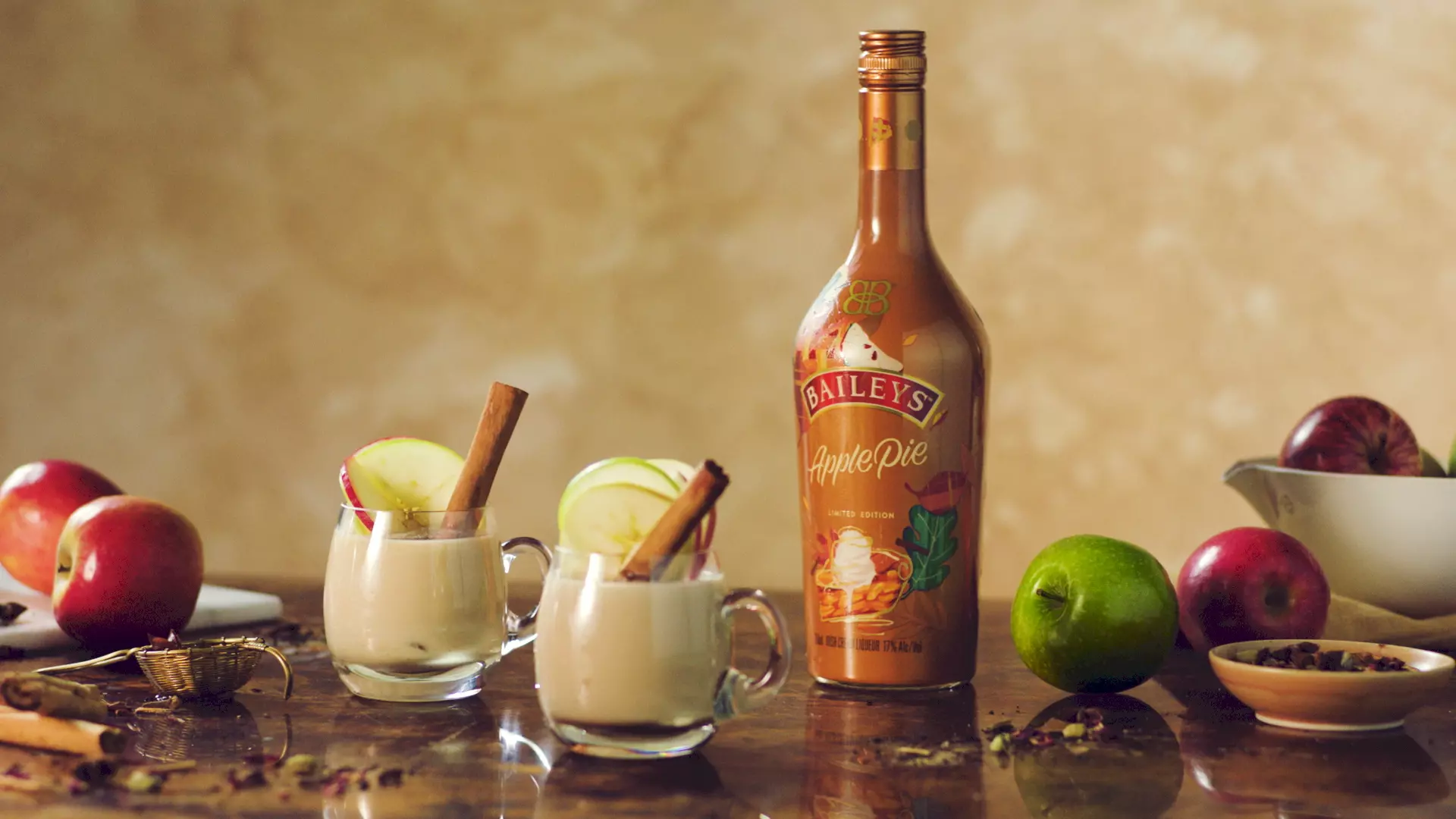 A bottle of Baileys Apple Pie liqueur stands next to two glass mugs of creamy drink garnished with apple slices and cinnamon sticks, surrounded by apples and spices on a wooden surface.