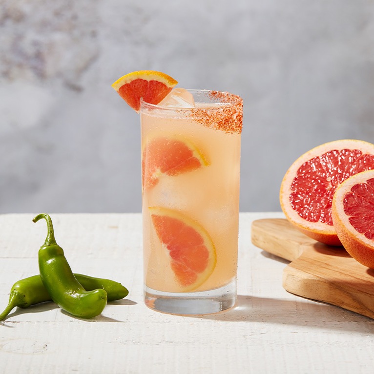 A Paloma cocktail next to a green chilli and a sliced grapefruit. 