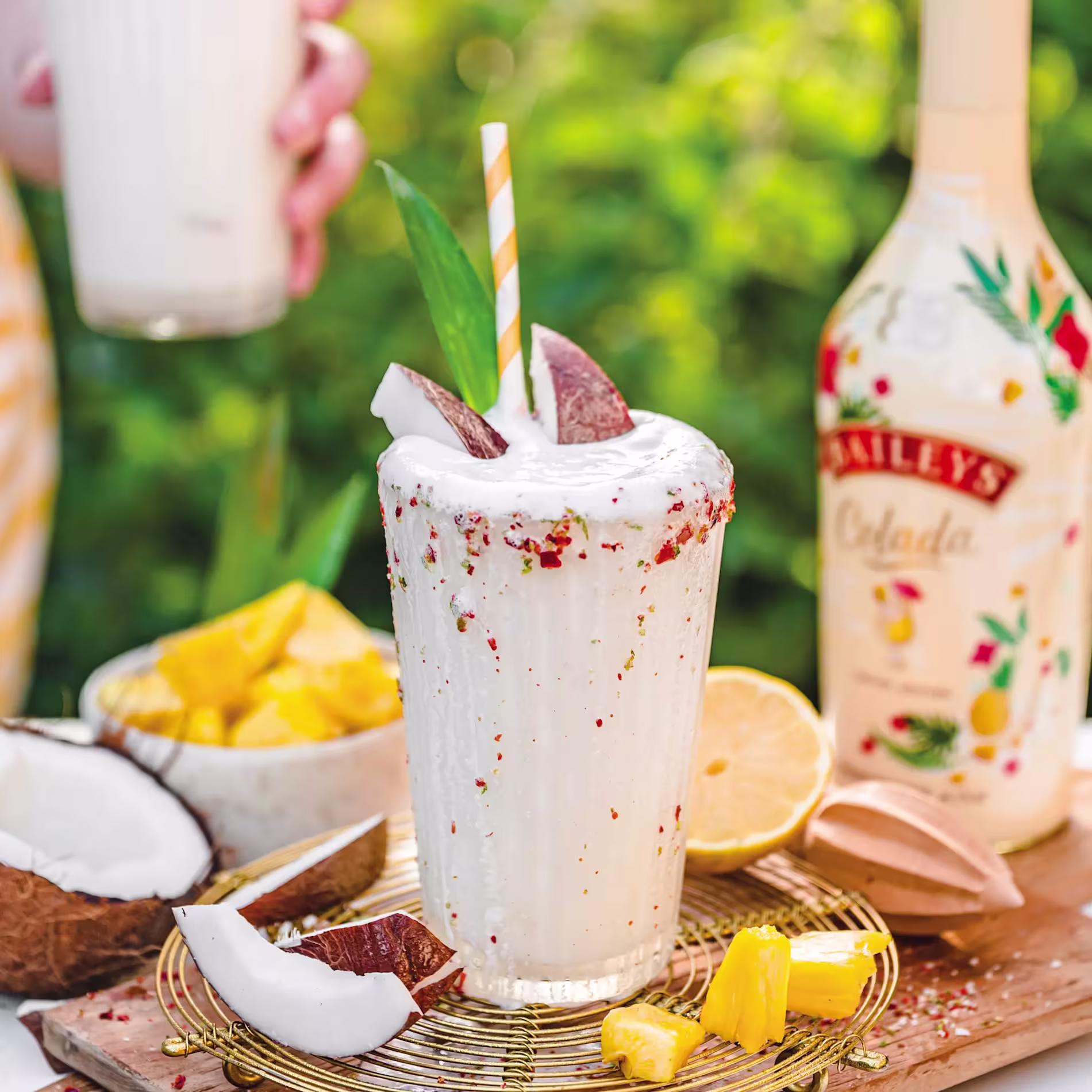 A tropical cocktail in a tall glass with a straw, garnished with coconut pieces, surrounded by pineapple chunks, coconut, lemon, and a bottle of Baileys Colada.