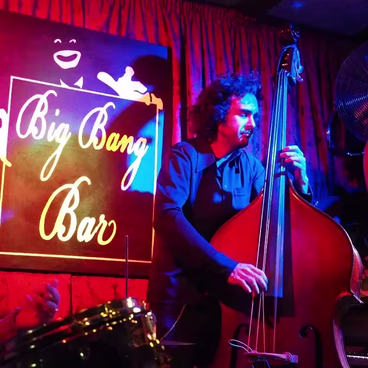 Músico tocando el contrabajo en el Big Bang Bar, iluminado por luces de colores frente al icónico cartel del local.