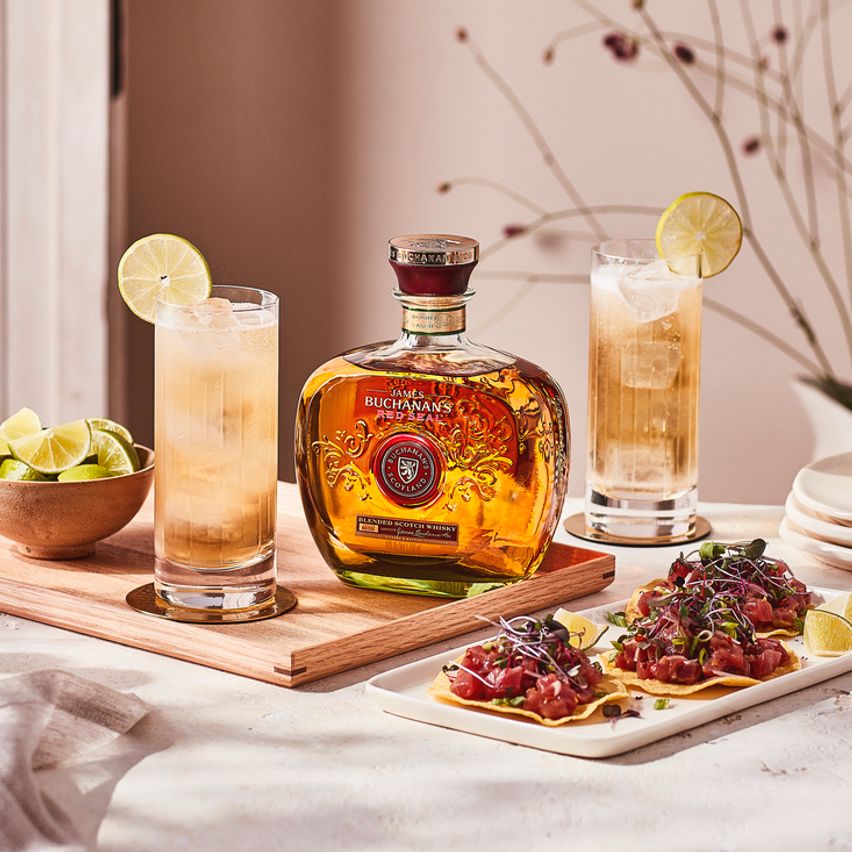 La imagen muestra una botella de whisky Buchanan's Red Seal junto a dos vasos con cócteles preparados con dicho destilado, acompañados de limones, y un plato de comida al costado.