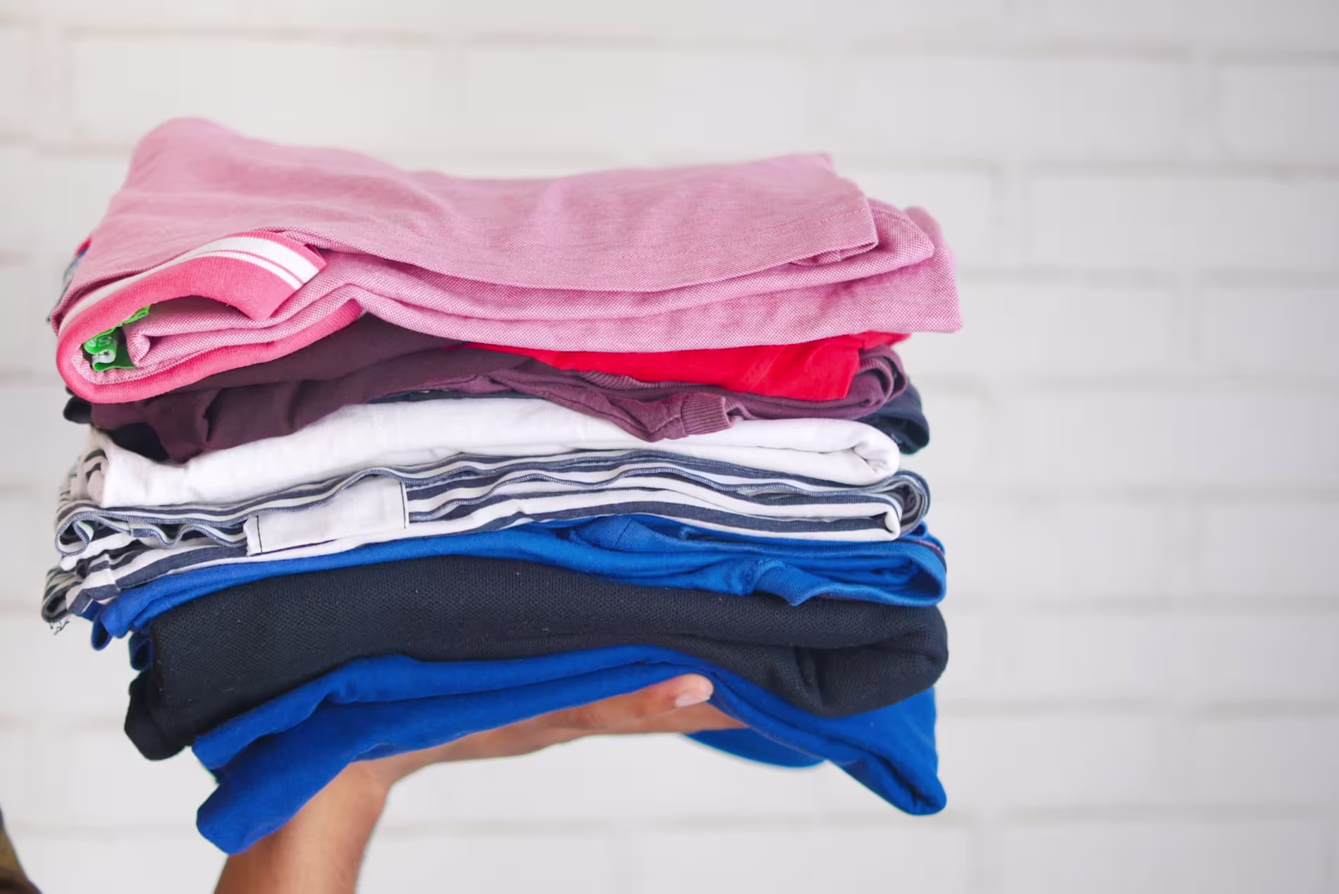A person holding a neatly folded stack of assorted clothes in various colors against a white brick background.