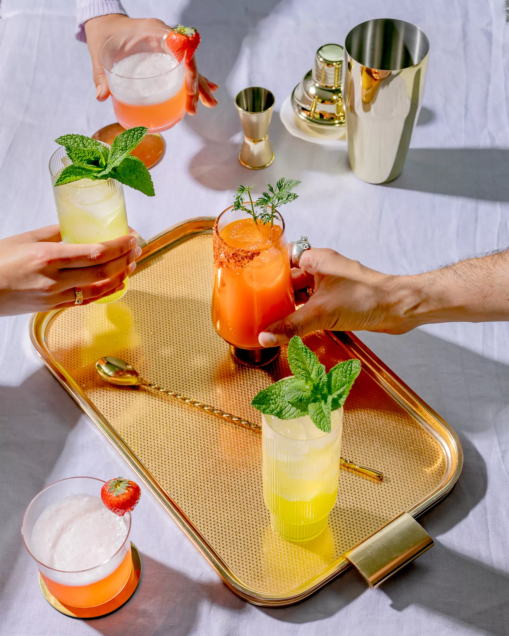 Three hands holding colorful cocktails over a gold tray with drinks, a spoon, and a cocktail shaker on a white tablecloth.