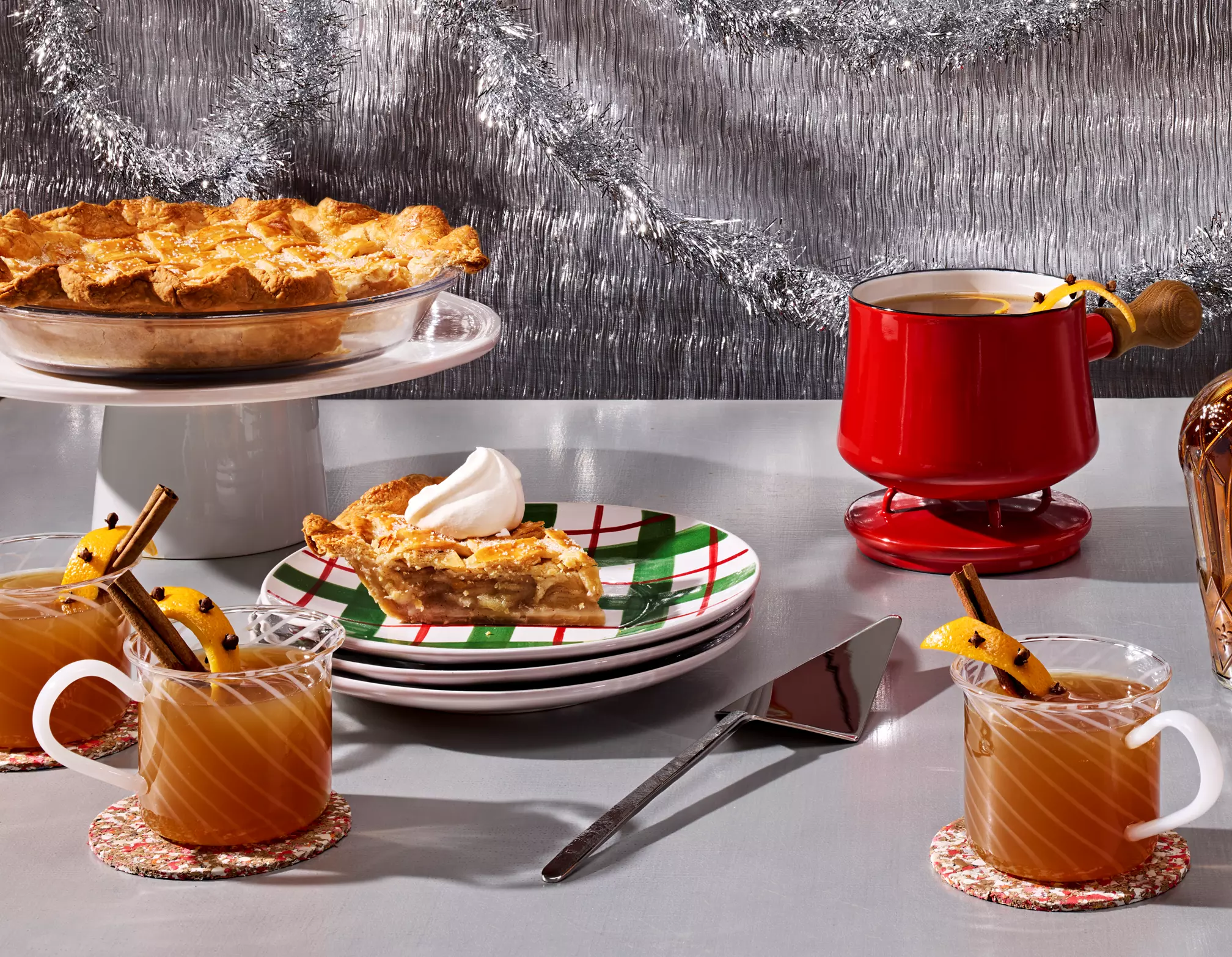A table with apple pie, a slice served on a plate with whipped cream, three mugs of spiced drink with cinnamon sticks, and a red mug, set against a silver tinsel backdrop.