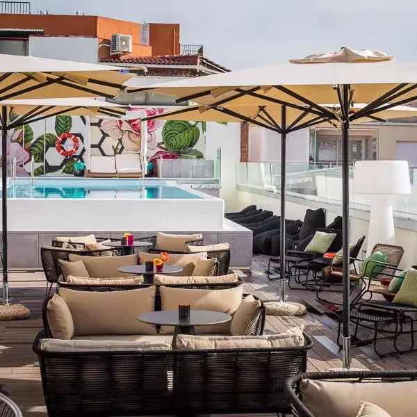 Terraza con piscina elevada, sofás con cojines claros, sombrillas y mesas bajas, rodeada de murales florales y edificios, ideal para relajarse al sol con estilo.