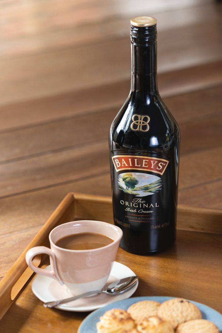 La imagen muestra una botella de Baileys Original Irish Cream, una taza rellena con Café Irlandés caliente con Baileys y un plato con galletitas dulces.