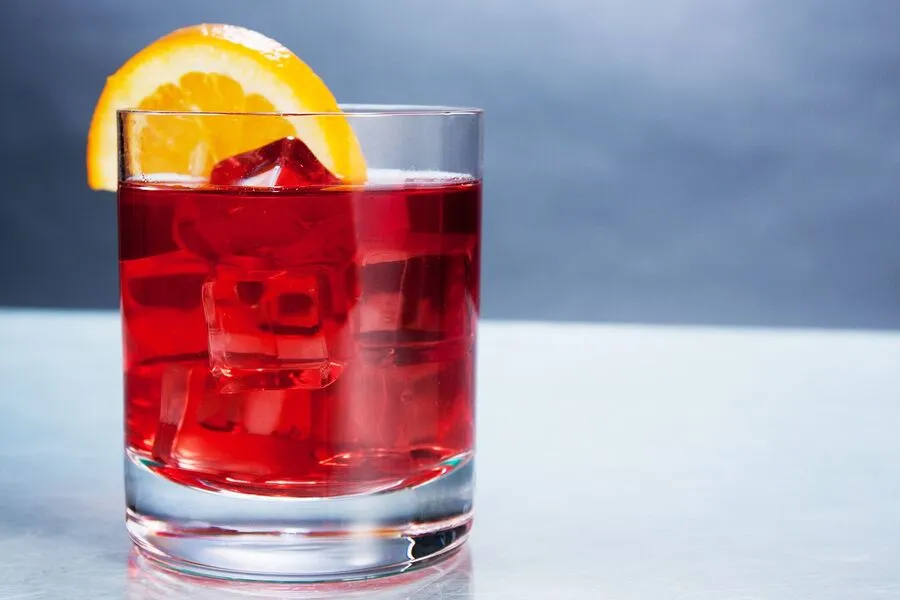 A clear glass filled with a red drink and ice cubes, garnished with a slice of orange on the rim, placed on a metallic surface.