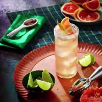 La imagen muestra un vaso largo relleno con el cóctel Paloma Don Julio y otros elementos de coctelería alrededor.