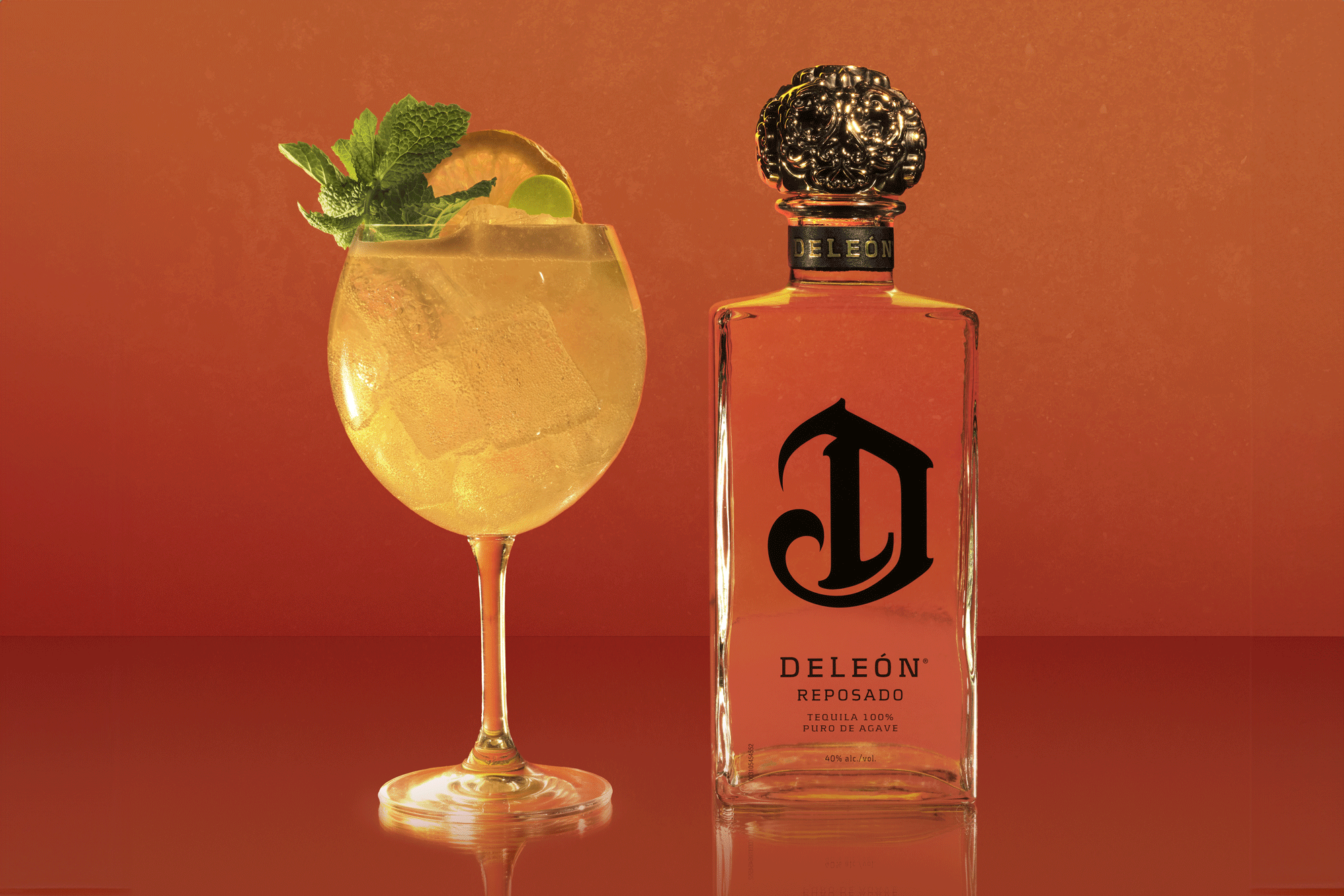 A glass of cocktail with ice, lime, and mint is placed next to a bottle of Deleón Reposado Tequila against an orange gradient background.