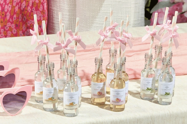 Glass bottles with pastel-colored drinks and pink bows on straws are arranged on a table next to heart-shaped sunglasses and a pink cloth.