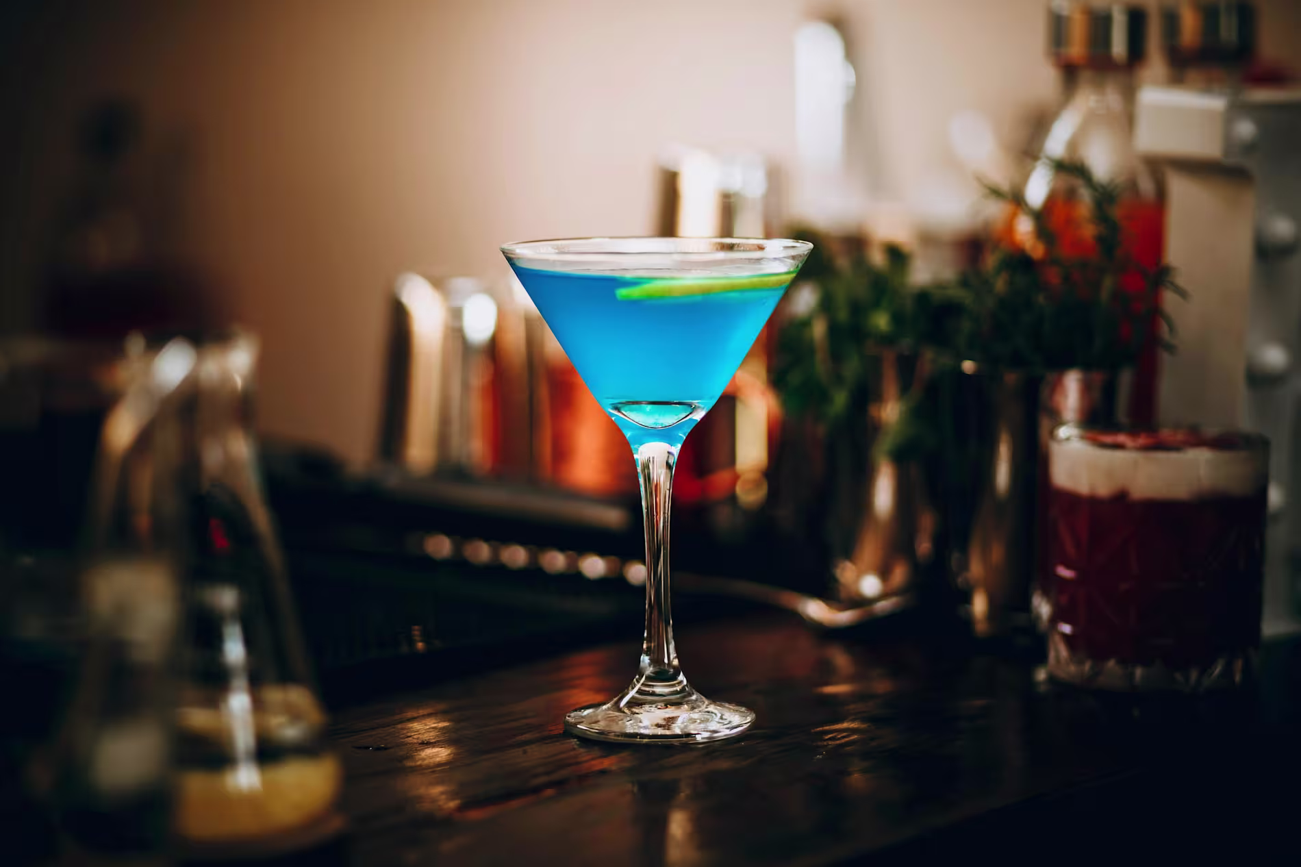 A blue cocktail in a martini glass garnished with a lime twist sits on a bar counter with bottles and shakers in the background.