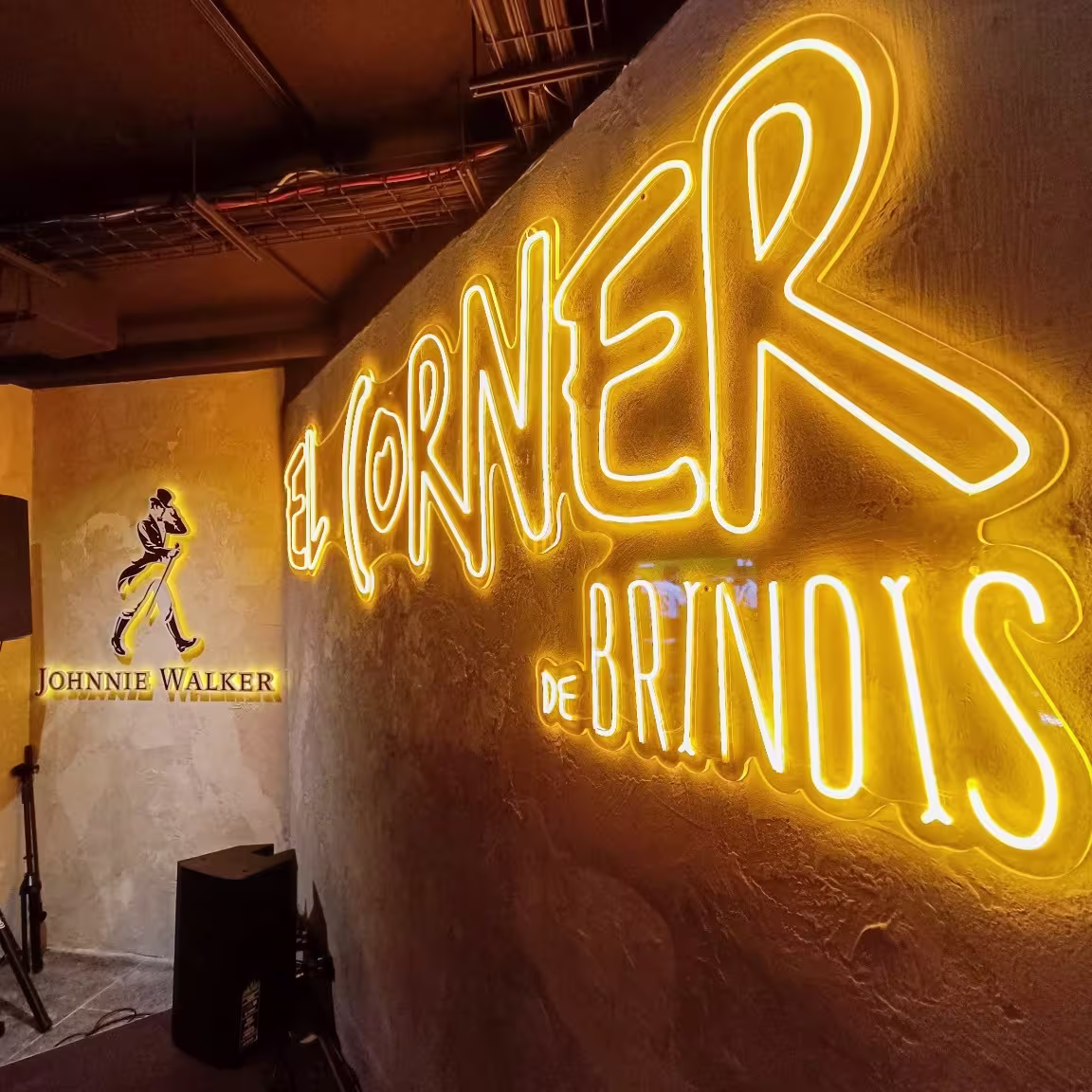 El Corner de Brindis: Entrada vibrante con neón amarillo resaltando el nombre del local y la marca Johnnie Walker en el fondo. Ambientes cálidos y modernos, ideales para momentos de celebración y brindis.