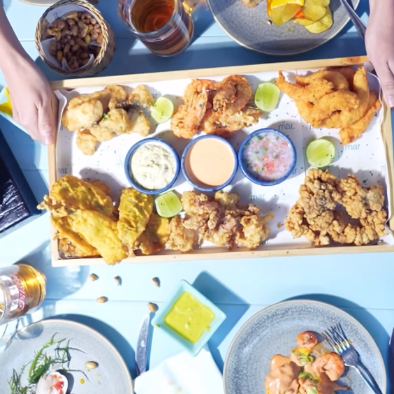 en la imagen se ve una mesa llena de comida y bebida: en el centro una bandeja con maricos fritos y sus respectivas salsas para acompañar