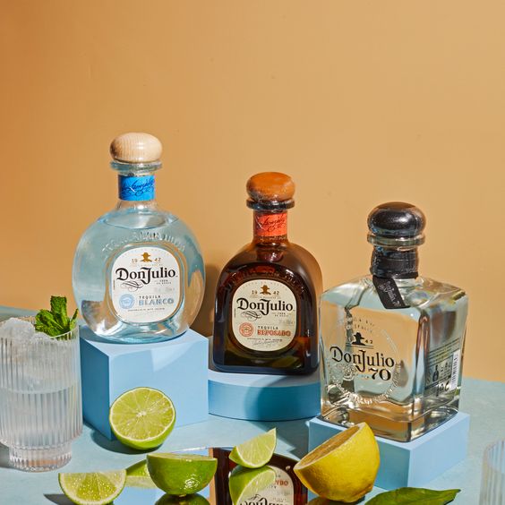 En la imagen se ven las botellas de Don Julio Reposado, Don Julio Blanco y Don Julio 70 sobre una tarima de color celeste. En el centro un espejo y limas y limones cortadas.