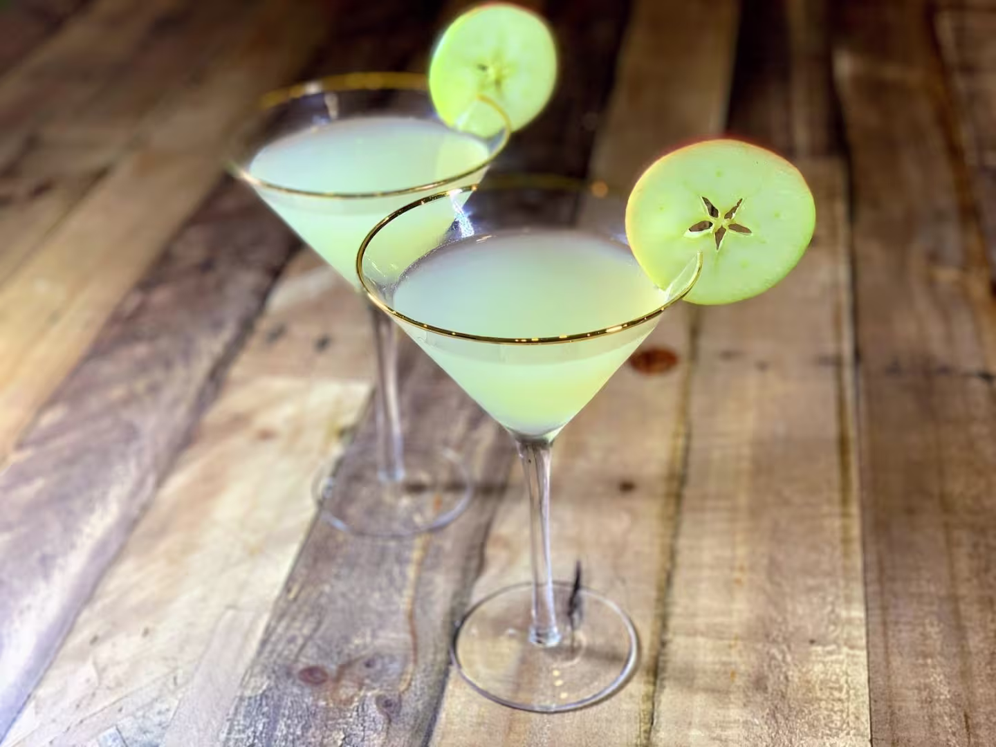 Two martini glasses filled with a pale green cocktail, each garnished with a slice of green apple, sit on a wooden surface.