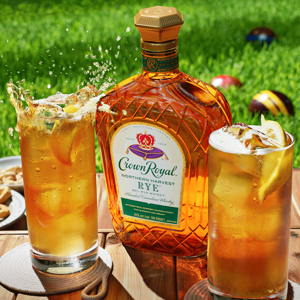 A bottle of Crown Royal Rye whiskey and two iced cocktails with lemon slices on a wooden table outdoors, with lawn games in the background.