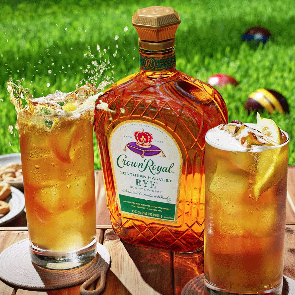 A bottle of Crown Royal Rye whiskey and two iced cocktails with lemon slices on a wooden table outdoors, with lawn games in the background.