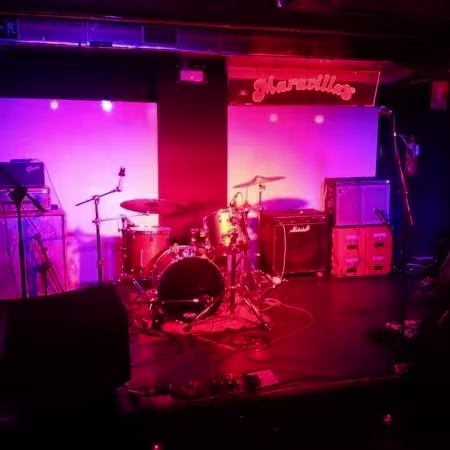 Maravillas Club: Escenario listo para el espectáculo, con luces en tonos morados y rojos que crean una atmósfera íntima y vibrante, rodeado de equipos de sonido y amplificadores que anticipan una noche de música en vivo.