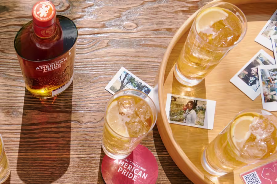 A bottle of American Pride whiskey, four cocktails with lemon slices, and several instant photos are arranged on a wooden surface and tray.