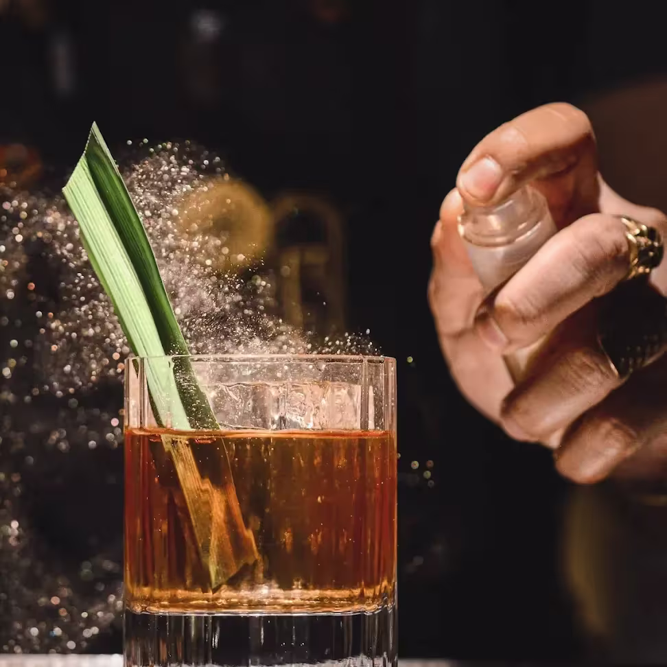 Cóctel en vaso bajo con hoja decorativa en Especiarium, mientras se rocía con un mist aromático que brilla bajo la luz.