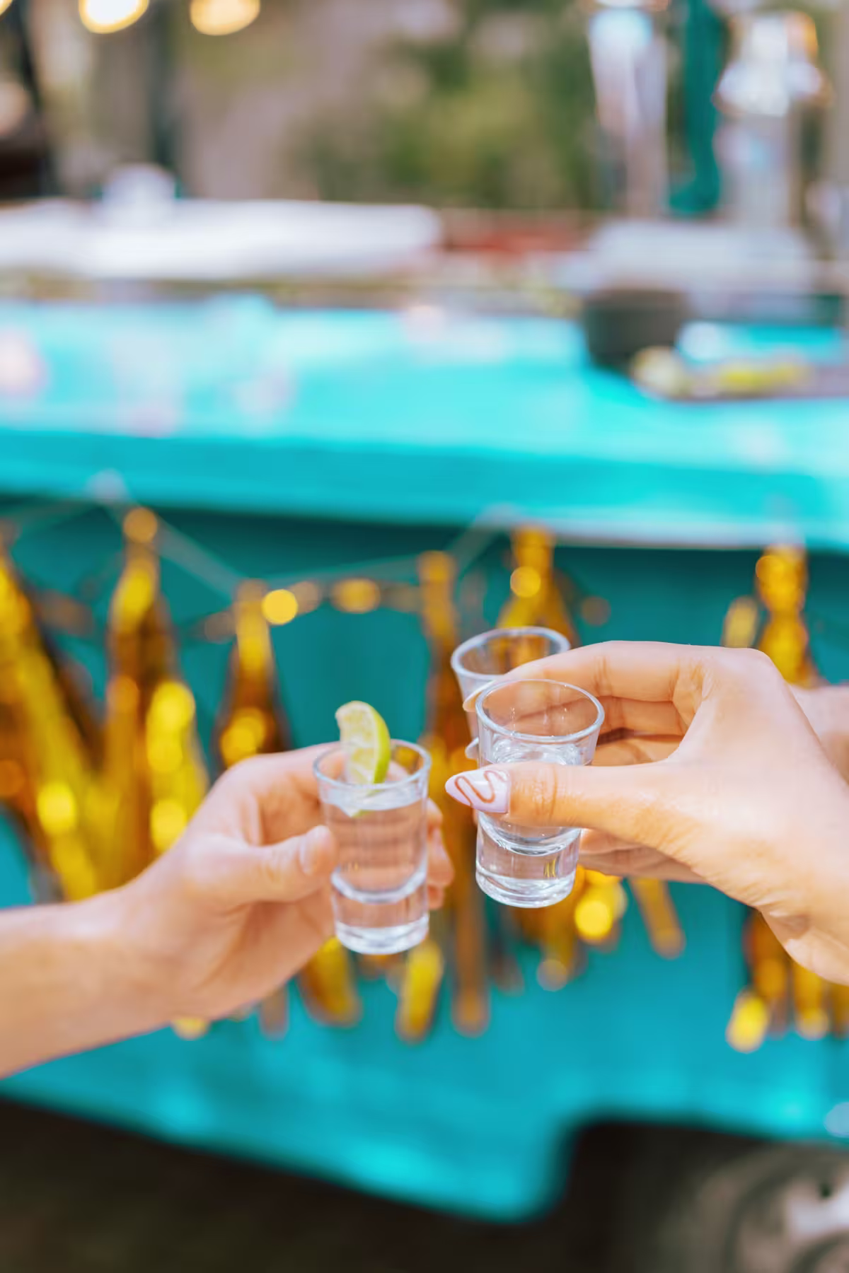 Three hands holding shot glasses in a toast, with one containing lime wedges, in front of a blue and gold festive background.
