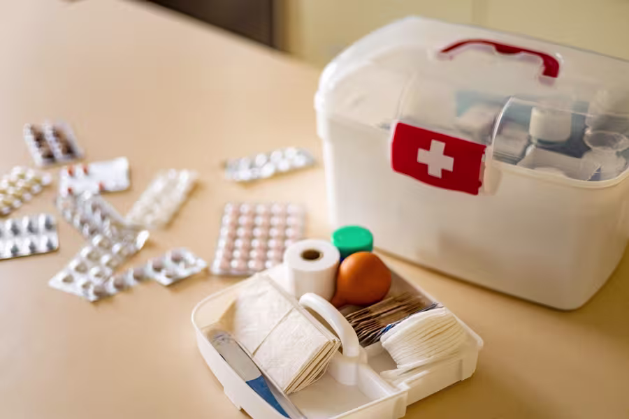 A first aid kit with bandages, gauze, tape, and ointments, with blister packs of pills and tablets spread on a beige surface nearby.