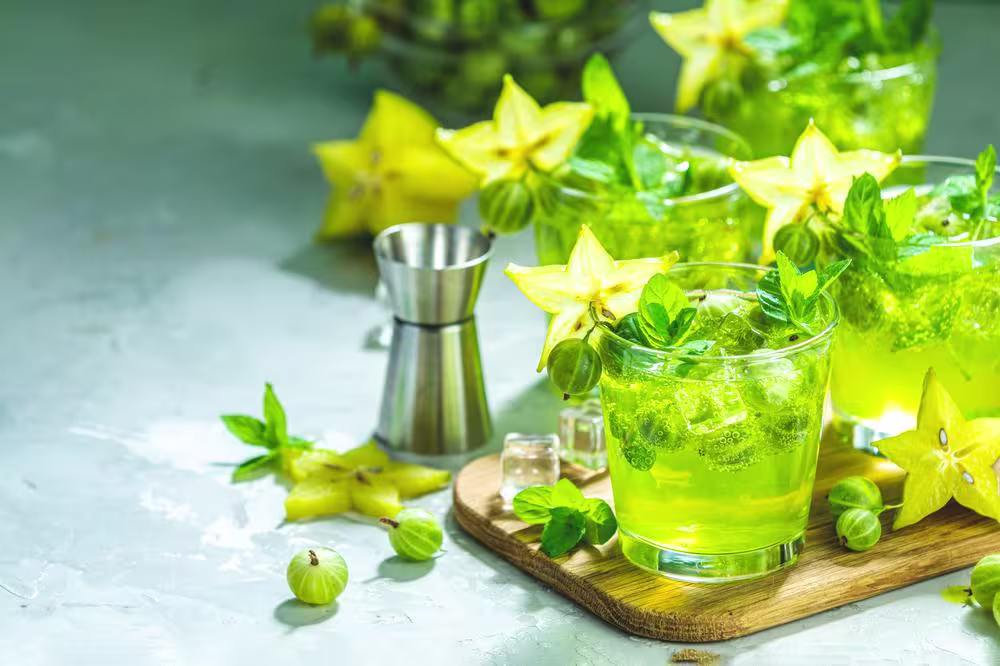 Glasses of green cocktail with mint, gooseberries, ice, and starfruit garnish sit on a wooden board beside a metal jigger and fresh starfruit slices.