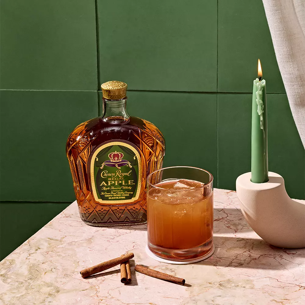 A bottle of Crown Royal Apple whisky, a glass of brown cocktail with ice, cinnamon sticks, and a green candle on a marble surface against a green tiled wall.