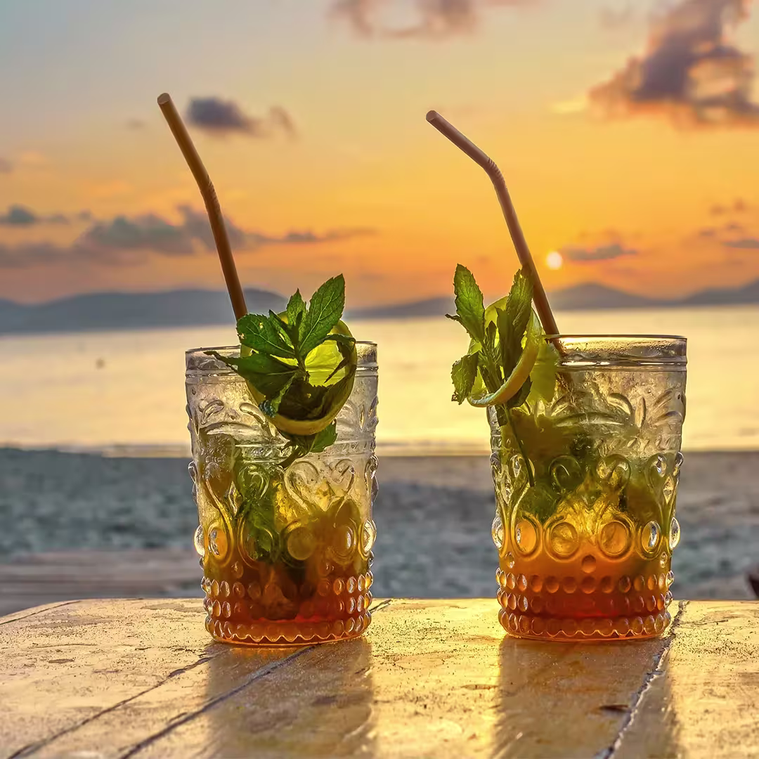Two cocktails with mint and lime sit on a table by the beach at sunset, with the ocean and an orange sky in the background.