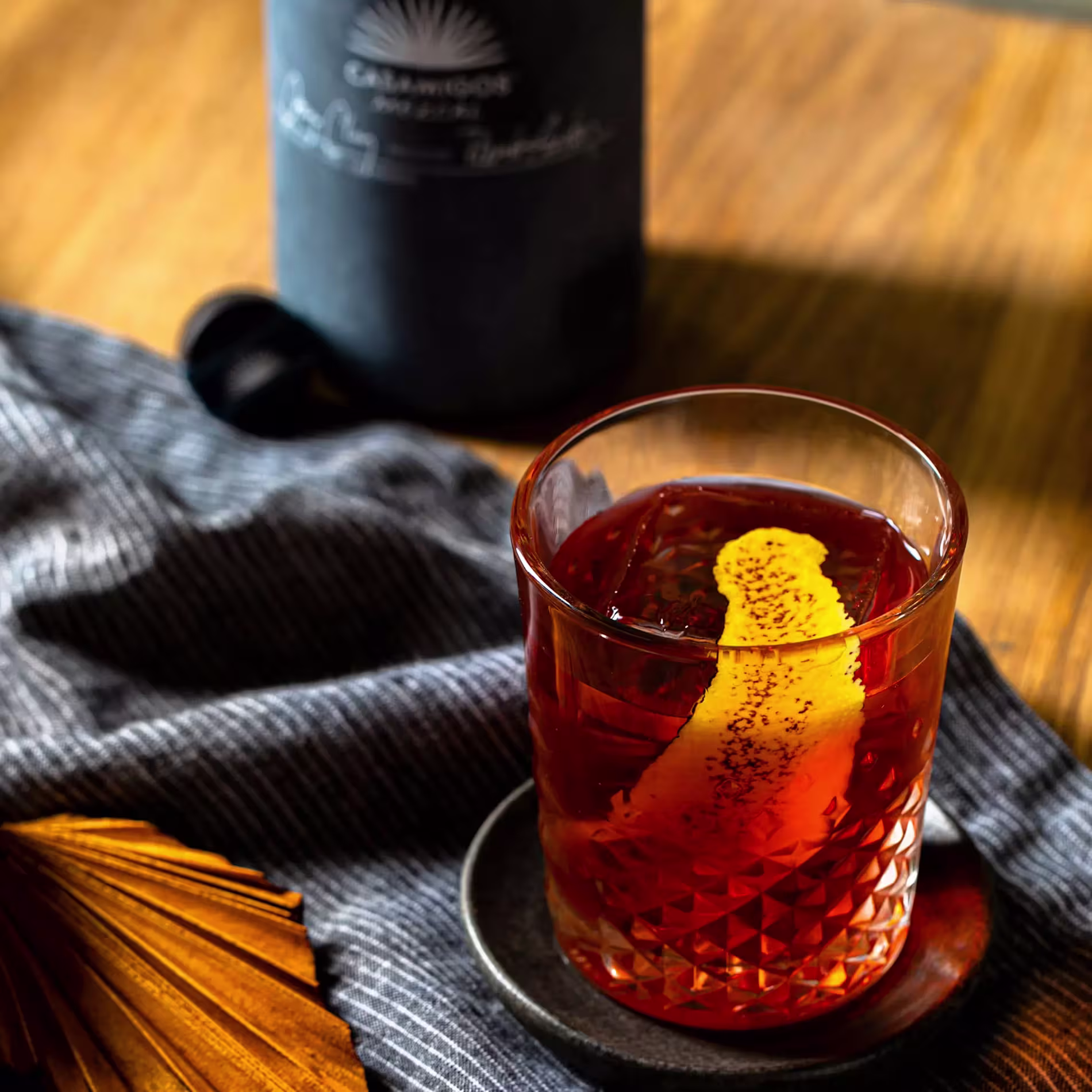A glass of amber cocktail with a citrus peel garnish sits on a small plate beside a striped cloth and a dark bottle in the background on a wooden surface.